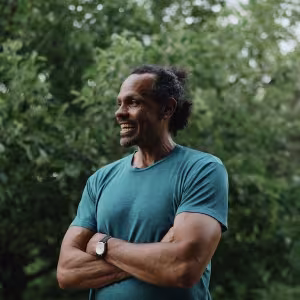 A dark-skinned man wearing a blue shirt stands with his arms crossed over his chest and a smile on his face in front of a backdrop of trees. 