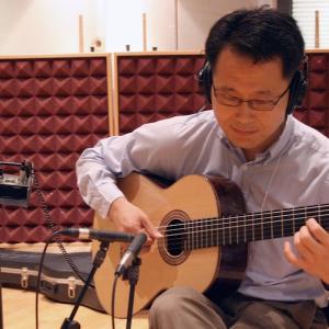A man wearing headphones and glasses plays a guitar in front of a microphone.