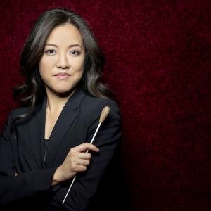 A dark-haired woman wearing a black suit and holding a conducting baton gazes at the viewer.