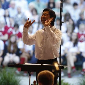 A conductor wearing a white shirt raises his hands and smiles at his orchestra.