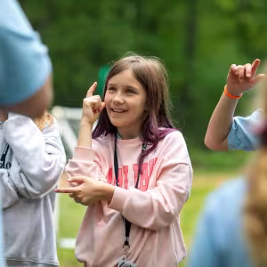 A student wearing a pink shirt smiles and lifts her arms as if dancing.