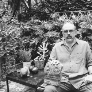 The artist wears a button-down shirt as he sits in a garden, holding a statue head on his lap. 