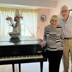 Two elderly individuals stand in front of a piano and smile.