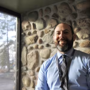 A bearded man with a tie stands in the light from a window and smiles.