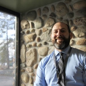 A bearded man with a tie stands in the light from a window and smiles.