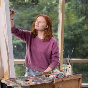A visual arts student with long red hair works on a painting. 