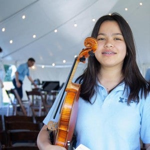 A young musician in a pale blue polo smiles as she holds her violin. 