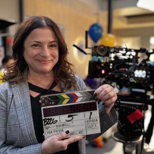 A smiling director poses with a clapperboard in front of camera equipment.