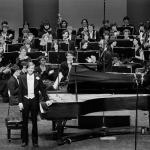 A pianist standing beside a grand piano in front of an orchestra in a black-and-white photo.
