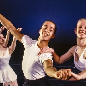 Male and female dancers smiling and holding hands in a staged performance with other dancers in the background.