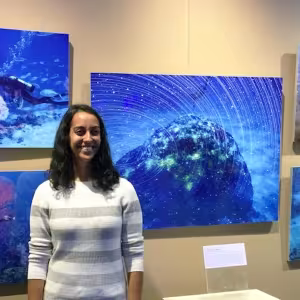  A woman in a striped sweater smiling at the camera in front of a wall displaying colorful underwater photographs of coral.