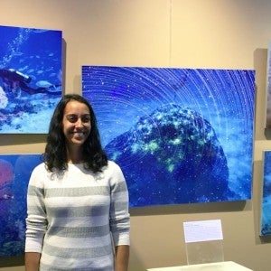  A woman in a striped sweater smiling at the camera in front of a wall displaying colorful underwater photographs of coral.