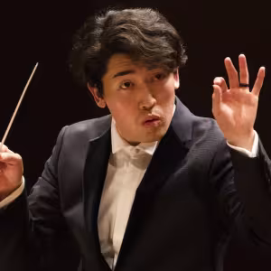 Person in a dark suit conducting with a baton, expressive gesture with the right hand, against a dark background.