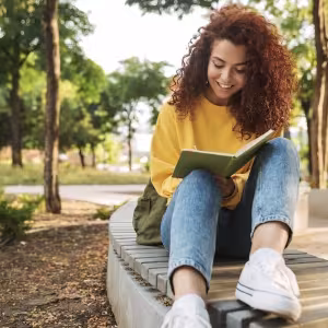 A student in a yellow sweater and jeans smiles as she reads a book.