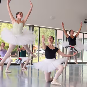 Four young dancers in tutus leap into the air, arms outstretched.