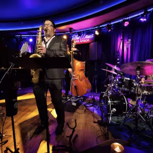 A saxophonist dressed in a dark suit plays passionately in front of a neon-lit stage with a drummer.