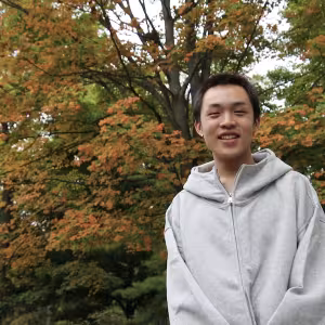 A student in a white hoodie stands against a fall landscape.