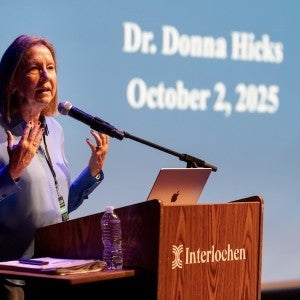 Dr. Donna Hicks, dressed in blue, gestures as she speaks from a podium.