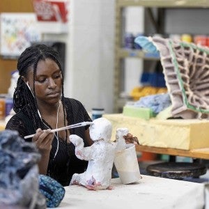 A seated student wearing earbuds paints a small white figure.