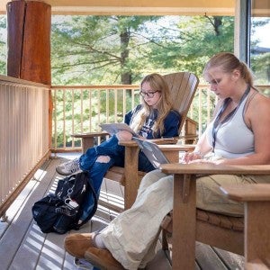 Two students sit comfortably on a deck as they type on their laptops.