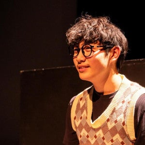 A student wearing an argyle vest sits in dramatic stage lighting.