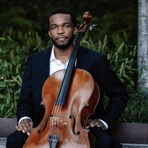 A musician wearing a suit sits with his cello against a natural backdrop.