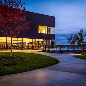 A large building with warmly-lit studio windows overlooks a shoreline at dusk.