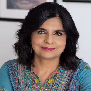 Professional headshot of a Parul Kapur with shoulder-length dark hair, wearing a teal embroidered top and earrings, smiling gently while facing the camera against a softly blurred indoor background with framed artwork.