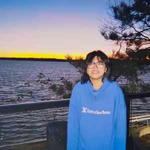 A student wearing a blue Interlochen hoodie poses against a lake at sunset. 
