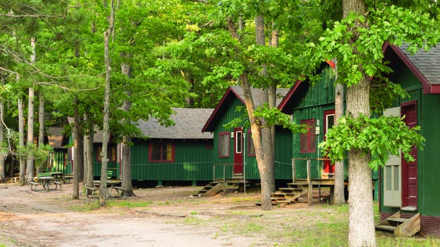 A row of dark green camp cabins with red doors and trim are pictured amongst dense trees in summer.