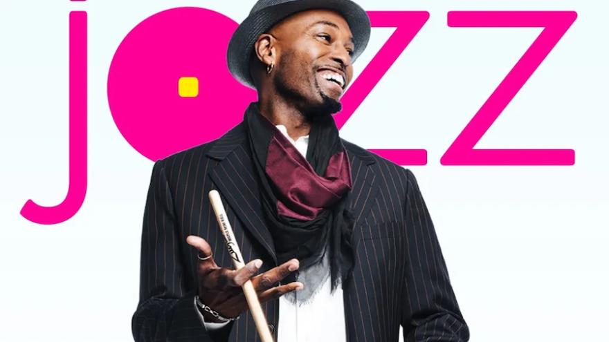 Clarence Penn , smiling, wearing a hat, and twirling drumstick , stands in front of hot pink lettering that reads "Jazz". 
